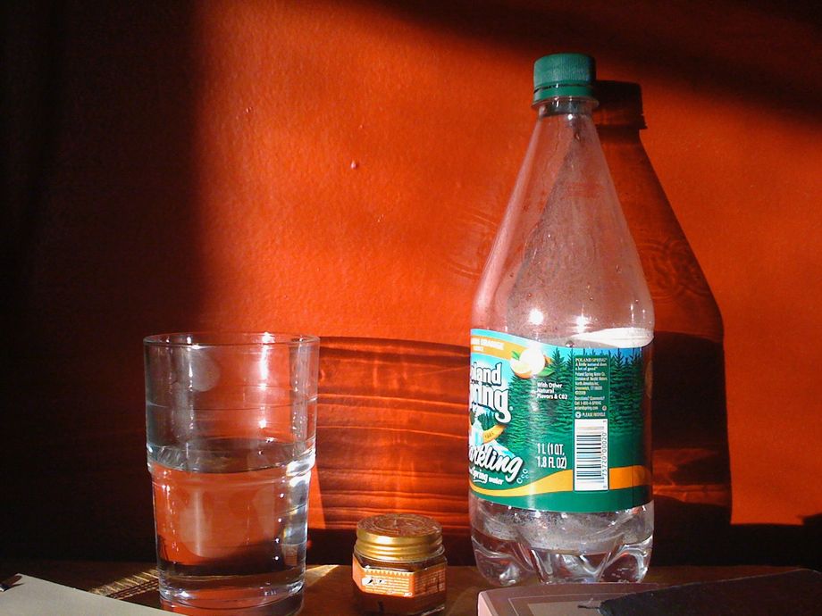 Early morning sun shines through a half-filled glass of water and a nearly empty water bottle, casting flat shadows on a deep red wall behind. Sleepy, peaceful. 