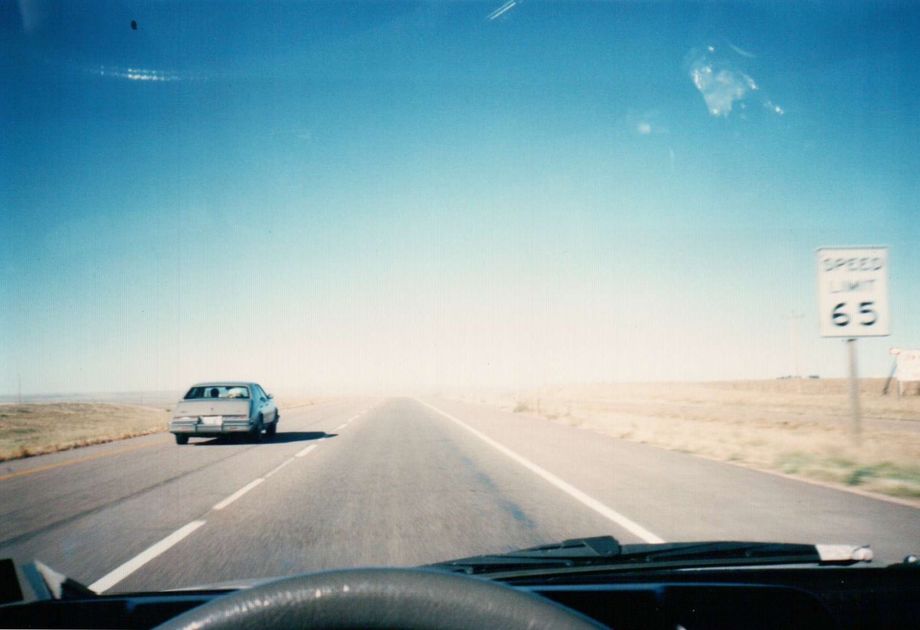 A grainy film photograph taken from the driver's seat of a vehicle in motion. The vehicle is in the right lane of a divided highway, being passed by another car to the left. To the right is a sign which reads 
