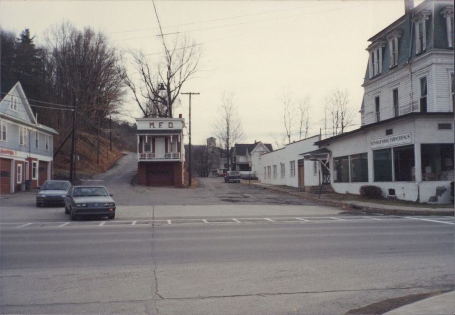 A film photograph of the intersections of Routes 6 and 26 in Meshoppen, Pennsylvania in February of 1987. The sky is vague and indistinct; the trees are bare, the film grain blurring the thin branches. A roadway crosses the bottom of the frame; two smaller roads curve away, one flat and level heading off into town, the other curving away to climb a wooded hill. Two dated looking cars are pulling out onto the highway. In between the two roads a sits a narrow, blocky building with a seemingly inaccessible second story porch and entryway. The wall above the short porch roof is white, and bears the handpainted block letters 