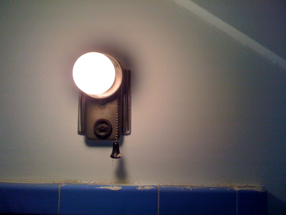 An antique wall outlet and light fixture houses a bare incandescent bulb, casting dim light and soft shadows. A short pull chain hangs from the fixture, the bell shaped end tapped lightly with paint matching the wall behind. The top of edge of tiles line the bottom of the frame, also streaked lightly with paint. A quiet and thoughtful solitude. 